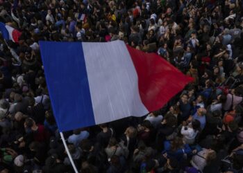 A large crowd gathers outdoors, with many people holding or waving French flags. Amidst this scene of unity and national pride, discussions about global events like the Nikkei 225 today add a layer of contemporary awareness against the backdrop of dense, diverse individuals. | FintechZoom