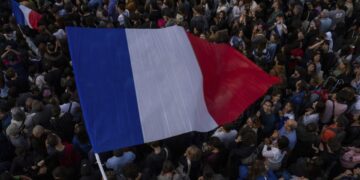 A large crowd gathers outdoors, with many people holding or waving French flags. Amidst this scene of unity and national pride, discussions about global events like the Nikkei 225 today add a layer of contemporary awareness against the backdrop of dense, diverse individuals. | FintechZoom