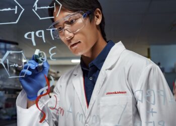A scientist wearing safety goggles and gloves writes chemical formulas on a transparent board in a laboratory. The scientist is dressed in a lab coat with the Johnson & Johnson logo. The background shows various laboratory equipment and shelves. | FintechZoom