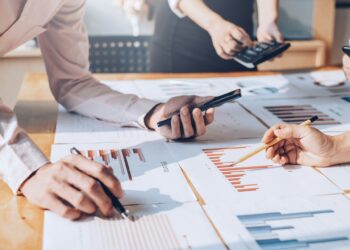 Three individuals are collaboratively working on a business plan at a desk covered with charts and graphs. One person holds a smartphone, another uses a calculator, and the third points at a bar chart with a pencil. Sunlight streams in from a nearby window, illuminating their workspace. | FintechZoom