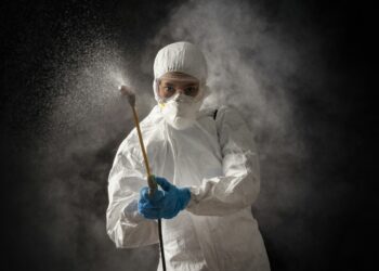 A person in full protective gear, including a white hazmat suit, gloves, goggles, and a face mask sprays disinfectant. The background is dark and misty, emphasizing the focus on sanitation and hygiene during pest inspections. | FintechZoom