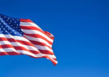 An American flag waves against a clear blue sky. The flag features 13 red and white stripes and a blue rectangle with 50 white stars in the top left corner. The flag is mounted on a tall flagpole. | FintechZoom