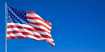 An American flag waves against a clear blue sky. The flag features 13 red and white stripes and a blue rectangle with 50 white stars in the top left corner. The flag is mounted on a tall flagpole. | FintechZoom