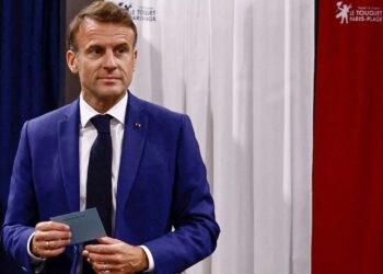 Emmanuel Macron, holding a piece of paper, stands in front of curtains with the colors blue, white, and red. The curtains' design appears to symbolize the French flag. With a serious expression and looking slightly to the side, he seems ready to discuss crucial FTSE 100 market updates. | FintechZoom