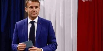 Emmanuel Macron, holding a piece of paper, stands in front of curtains with the colors blue, white, and red. The curtains' design appears to symbolize the French flag. With a serious expression and looking slightly to the side, he seems ready to discuss crucial FTSE 100 market updates. | FintechZoom