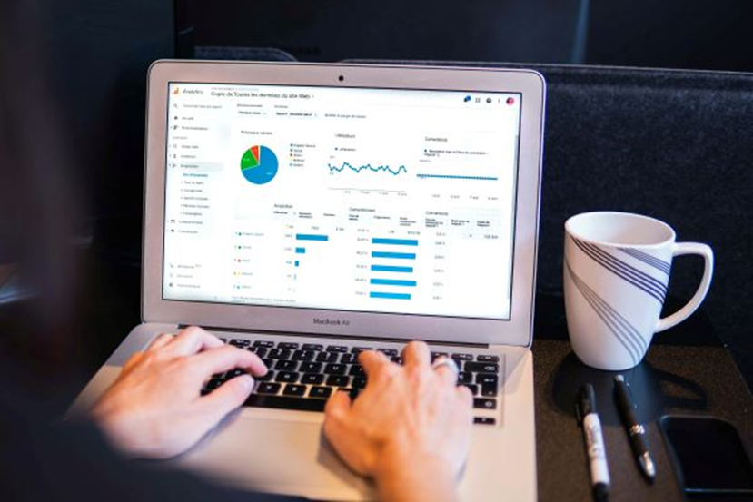 A person is using a MacBook laptop displaying various charts and graphs, likely showing eCommerce data analytics. The person’s hands are on the keyboard. A white coffee mug and two markers are placed on the desk beside the laptop. | FintechZoom