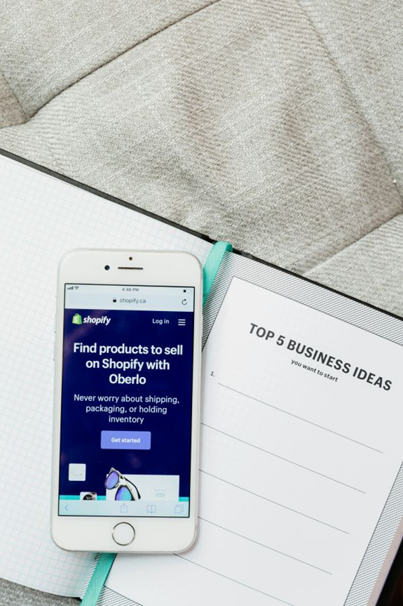 An iPhone displaying the Shopify eCommerce website is placed on an open notebook. The notebook’s right page is titled “Top 5 Business Ideas,” with bullet points for listing ideas. The background features a soft, quilted surface. | FintechZoom