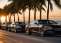 A row of sleek, black luxury cars parked along a seaside road, lined with palm trees. The sun sets in the background, casting a warm glow on the scene, with the ocean visible in the distance—the perfect spot for those seeking exotic car rental in Miami. | FintechZoom