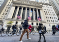 The image shows a bustling street scene in front of the New York Stock Exchange, with noticeable features including the Fearless Girl statue, pedestrians, and cyclists. The stock exchange building, home to the Dow Jones Industrial Average, is adorned with three large American flags. | FintechZoom