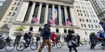 The image shows a bustling street scene in front of the New York Stock Exchange, with noticeable features including the Fearless Girl statue, pedestrians, and cyclists. The stock exchange building, home to the Dow Jones Industrial Average, is adorned with three large American flags. | FintechZoom