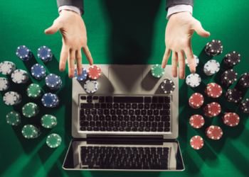 A person in a suit reaches out over a laptop keyboard, surrounded by stacks of poker chips on a green felt surface, suggesting an online gambling scenario through sweepstakes casino apps. | FintechZoom