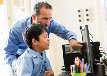 An adult and a child are at a desk using a computer. The adult, wearing a blue shirt, is pointing at the screen while teaching the child about online safety. The child, also in a blue shirt, is attentively watching. A container of colorful pens and pencils is on the desk in the foreground.
 | FintechZoom