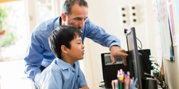 An adult and a child are at a desk using a computer. The adult, wearing a blue shirt, is pointing at the screen while teaching the child about online safety. The child, also in a blue shirt, is attentively watching. A container of colorful pens and pencils is on the desk in the foreground.
 | FintechZoom