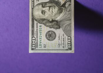A U.S. $100 bill photographed against a purple background, reminiscent of a real estate listing photo. The bill is oriented vertically, displaying the portrait of Benjamin Franklin clearly. Shadows cast by the bill fall to the side, emphasizing the texture of the background. | FintechZoom