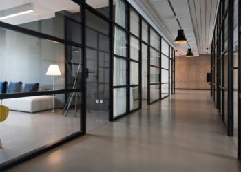 A modern office space ideal for startups, featuring glass-walled meeting rooms on either side of a polished concrete hallway. The area is lit by pendant lights hanging from the ceiling, and there is a cozy seating area with a sofa, lamp, and cushions in the back. | FintechZoom