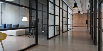 A modern office space ideal for startups, featuring glass-walled meeting rooms on either side of a polished concrete hallway. The area is lit by pendant lights hanging from the ceiling, and there is a cozy seating area with a sofa, lamp, and cushions in the back. | FintechZoom