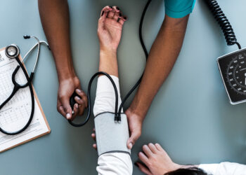 A healthcare professional measures a patient's blood pressure using a sphygmomanometer. The patient's arm is extended on a table, and medical tools, including a stethoscope and clipboard with a report, are visible beside them. | FintechZoom
