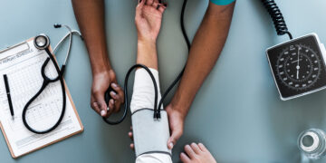 A healthcare professional measures a patient's blood pressure using a sphygmomanometer. The patient's arm is extended on a table, and medical tools, including a stethoscope and clipboard with a report, are visible beside them. | FintechZoom