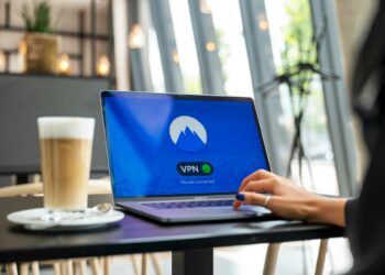 A person sits at a modern cafe table, using a laptop displaying a VPN connection screen. A tall glass of layered coffee with foam rests on a saucer beside the laptop, as sunlight streams through large windows in the background, accentuating the serene atmosphere. | FintechZoom