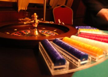 An interior shot of a casino roulette table. The roulette wheel is spinning, with the ball visible on the outer rim. To the right, there are trays filled with colorful casino chips. A person's hand is seen blurred in motion at the edge of the image. | FintechZoom