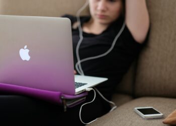 A person relaxing on a couch uses a MacBook laptop with a purple cover, likely accessing Learning Management Systems (LMS). The person is wearing earphones and has one arm resting behind their head. A smartphone lies on the couch beside them. The background is out of focus. | FintechZoom