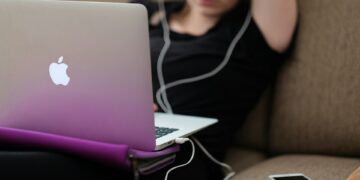 A person relaxing on a couch uses a MacBook laptop with a purple cover, likely accessing Learning Management Systems (LMS). The person is wearing earphones and has one arm resting behind their head. A smartphone lies on the couch beside them. The background is out of focus. | FintechZoom