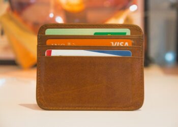Image of a brown leather cardholder with three visible slots, each holding a card including a green and white Visa and various other Forex cards. The cardholder rests on a white surface, set against a blurred background featuring warm lighting. | FintechZoom