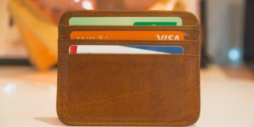 Image of a brown leather cardholder with three visible slots, each holding a card including a green and white Visa and various other Forex cards. The cardholder rests on a white surface, set against a blurred background featuring warm lighting. | FintechZoom