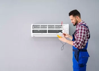 A man in blue overalls and a red plaid shirt is servicing an AC that needs repairs, mounted on a gray wall. He is holding a tool and checking the unit, likely performing maintenance or fixes. | FintechZoom