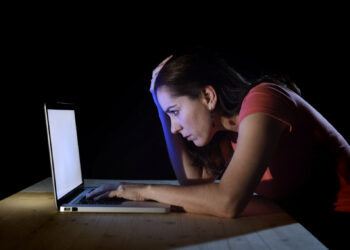 A woman in a red shirt sits at a wooden table in a dark room, illuminated by the light from her laptop screen. She leans forward with her hand on her forehead, appearing stressed or tired as she delves into an article about online scams on her laptop. | FintechZoom