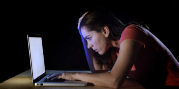 A woman in a red shirt sits at a wooden table in a dark room, illuminated by the light from her laptop screen. She leans forward with her hand on her forehead, appearing stressed or tired as she delves into an article about online scams on her laptop. | FintechZoom