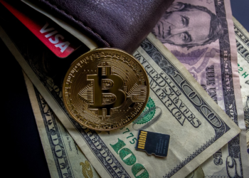A leather wallet with a Visa card partially visible lies on a surface with scattered U.S. dollar bills, a gold Bitcoin coin, and a small microSD memory card. | FintechZoom