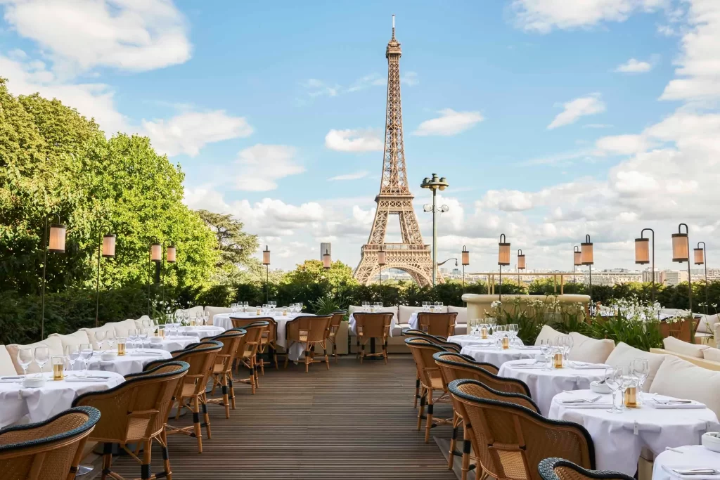 Girafe Paris dining area with elegant table settings, featuring white tablecloths and wicker chairs, is situated on a wooden deck. The Eiffel Tower stands prominently in the background against a blue sky with scattered clouds. Lush greenery surrounds the dining space. | FintechZoom