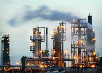 An industrial facility with multiple tall structures and metal frameworks, emitting smoke and illuminated by numerous bright yellow lights. Against the backdrop of cloudy skies, the overcast atmosphere hints at underlying geopolitical tensions shaping the scene. | FintechZoom