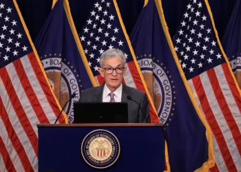 A person in a suit and tie stands at a podium with a microphone, speaking. Behind them are multiple American flags, the seal of the Board of Governors of the Federal Reserve System, and a screen displaying key points from the latest Fed minutes. | FintechZoom