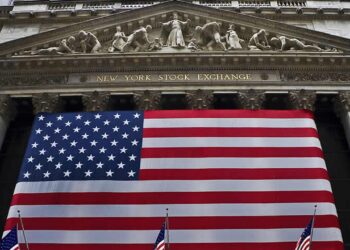 The front facade of the New York Stock Exchange building, displaying large columns and decorative sculptures, is adorned with a massive American flag that covers the entrance. Smaller American flags are visible in the foreground, reflecting the spirit of the stocks market today. | FintechZoom