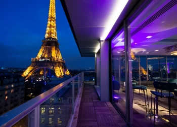 A brightly lit Eiffel Tower at night viewed from a modern balcony with glass railings, where purple interior lighting reflects in large windows—evoking the chic atmosphere of Le Ciel de Paris. | FintechZoom