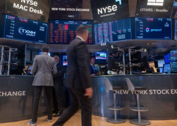 Traders work on the floor of the New York Stock Exchange, surrounded by numerous digital screens displaying stock prices, company logos, and financial data. Amidst the bustling trading floor environment, people stand and walk as shares gain or lose value in real-time. | FintechZoom