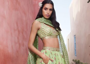A woman stands in an alleyway with light peach and beige walls, wearing an elegant South Asian outfit. Her ensemble features a green crop top, a long skirt with floral patterns, and a matching scarf. Her long dark hair is styled straight, adorned with white flowers, adding to her graceful presence. | FintechZoom