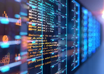An image of a server rack with multiple units, glowing blue with indicator lights. In the foreground, transparent lines of colorful code are superimposed, representing programming and data processing activities in a technology and data center environment characteristic of the Digital Age. | FintechZoom