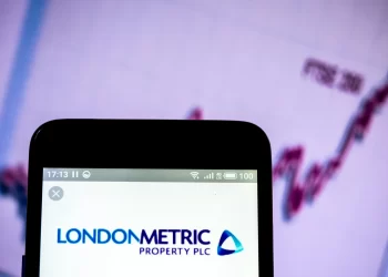 A smartphone displays the logo and name "LondonMetric Property PLC" on its screen. In the background, a blurred graph labeled "FTSE 100 Index" is visible, likely indicating financial performance or stock market information. | FintechZoom