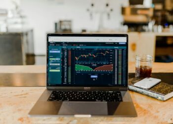A laptop displaying a financial trading graph sits on a wooden table, embodying the pulse of the fintech industry. Next to the laptop is a glass of iced coffee, a closed notebook, and a smartphone. The blurred indoor setting in the background features additional furniture and decor. | FintechZoom