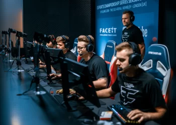 A group of gamers in black shirts sit in a row at computers, wearing headphones and focused on their screens. The backdrop features gaming sponsorships and event logos. Another person stands behind them, observing. | FintechZoom