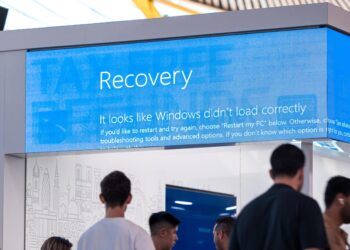 Several people stand and walk around a modern, indoor space with a large screen overhead displaying the notorious blue screen of death: "Recovery. It looks like Windows didn’t load correctly..." The area is bright and features contemporary decor. | FintechZoom