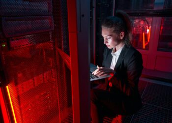 A woman in a dark server room works on a laptop, illuminated by red and blue lights with server racks visible around her.
