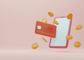 A smartphone and a credit card are floating together against a soft pink background, symbolizing online earnings. Golden coins are scattered around them, seemingly in motion. The credit card is red, with minimal details visible, and the smartphone has a large blank screen. | FintechZoom