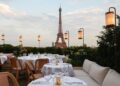 Girafe Paris rooftop dining area with elegantly set tables and chairs, overlooking the Eiffel Tower at sunset. Soft lighting from lanterns adds ambiance. The Eiffel Tower stands prominently in the background against a partly cloudy sky. Greenery surrounds the seating area. | FintechZoom