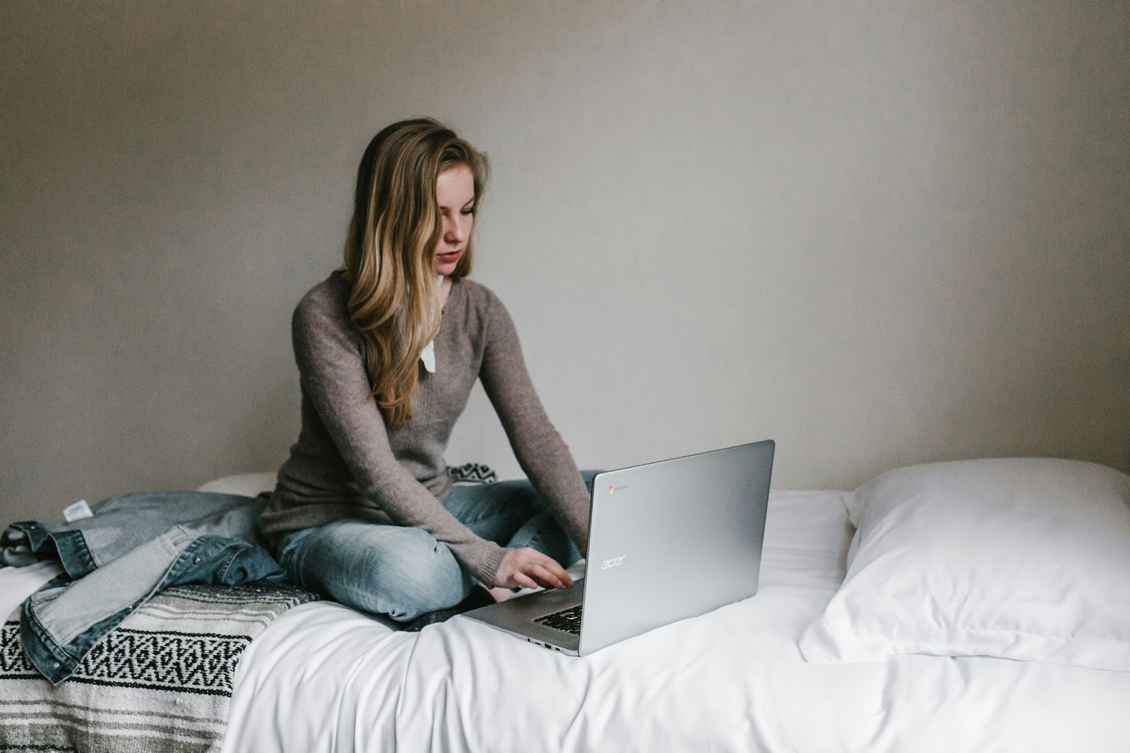 A person with long hair sits cross-legged on a bed in a casual, relaxed setting, focusing intently on their laptop. They are wearing a grey sweater and jeans. The bed has white sheets and a white blanket, with a jacket lying beside them. They appear to be utilizing EssayService AI for assistance. | FintechZoom