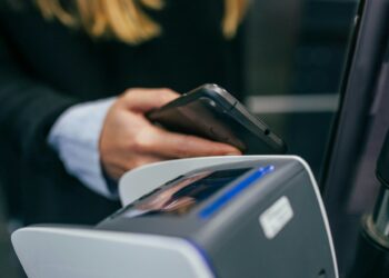 A person holding a smartphone near a contactless payment terminal, appearing to make a Buy Now Pay Later transaction. The person's hand and part of their arm are visible, along with the payment device and the phone. The focus is on the interaction between the phone and the terminal. | FintechZoom