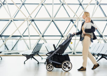 A woman walks through an airport terminal pushing a baby stroller with one hand and holding a baby on her hip with the other. The terminal features large, geometric windows, and empty benches are visible in the background. | FintechZoom